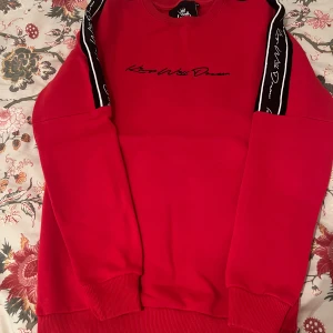 Röd sweatshirt från Kings Will Dream XS - Röd sweatshirt från KWD i strl XS med svarta och vita detaljer längs ärmarna och tryck på bröstet. Baksidan har stora svarta ränder och text.  Perfekt för en streetwear-look. Tröjan är bara använd 1 gång.