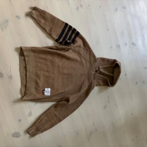 Beige hoodie med ränder - Säljer en beige stickad hoodie liknande Thom Browne med fyra svarta ränder på ena ärmen. Tröjan har huva med snörning och ribbade muddar. Perfekt för dig som gillar stilrena detaljer och exklusiva kläder