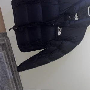 Svart dunjacka från The North Face 1996 - Säljer en svart dunjacka från The North Face med klassisk logga på bröstet. Jackan har hög krage, dragkedja och är riktigt puffig och varm. Perfekt för kalla dagar och snygg till streetwear-stilen. Använt lite 