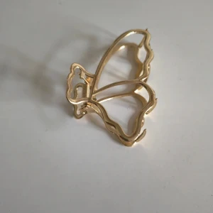 Guldig fjärilsformad hårklämma - En snygg hårklämma i metall med fjärilsformad design och guldig finish. Klämman har en öppen, stilren look och passar perfekt för att sätta upp håret på ett trendigt sätt. Enkel att använda och ger en extra touch till din frisyr. Aldrig använd.