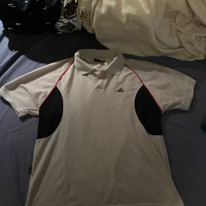 Beige pikétröja  - Snygg beige pikétröja från Rough med svarta paneler på sidorna och röda detaljer längs axlarna. Klassisk krage och korta ärmar, perfekt för en sportig look. Broderad logga på bröstet. Skön och luftig bomullskvalitet.