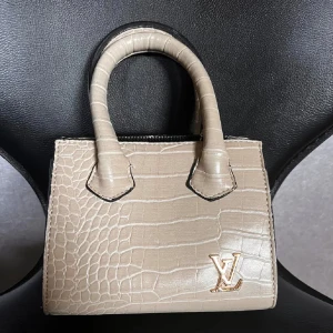 Beige handväska  - Snygg beige handväska INTE ÄKTA!!!!, med krokopräglat mönster och guldfärgad  logga. Väskan har två handtag och en avtagbar kedja i guld, perfekt för att bära både i handen och över axeln. Stilren och lyxig design som lyfter varje outfit.