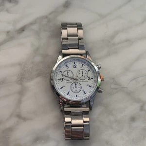 Silverfärgad kronografklocka PRC 200 - Stilren kronografklocka i silverfärgat metallarmband och vit urtavla. Klockan har tre mindre urtavlor, tydliga siffror och tachymeter runt kanten. Metallarmbandet ger en klassisk och clean look, perfekt för dig som gillar detaljer och sportig design.