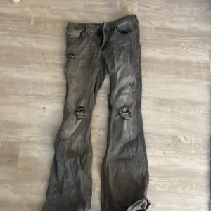 Grå slitna bootcut jeans 157 Lowboot - Grå jeans från 157 i modellen Lowboot med bootcut passform. Jeansen har slitningar och hål på båda benen för en riktigt edgy look. Tillverkade i klassiskt jeanstyg och har normal längd. Perfekta för dig som gillar en avslappnad och trendig stil. ”Hållen är gjorda själv” och passar storlek s