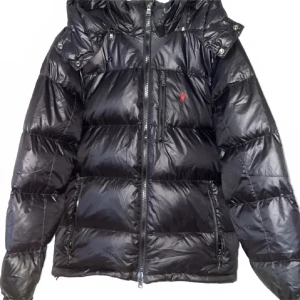 Svart pufferjacka Polo Ralph Lauren S - Svart pufferjacka från Polo Ralph Lauren med glansig finish, huva och dragkedjor framtill. Klassisk röd logga på bröstet och flera praktiska fickor. Perfekt för kalla dagar när du vill hålla stilen och värmen.