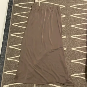 Brun långkjol i satin - Säljer en stilren brun långkjol i satin med mjuk glansig yta och enkel design. Kjolen har resår i midjan och faller snyggt längs benen. Perfekt för dig som gillar minimalistisk och trendig stil. Från VILA