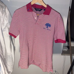 Randig pikétröja Polo Golf röd/vit - Snygg randig pikétröja från Polo Golf i rött, vitt och blått. Tröjan har klassisk krage, korta ärmar och knappar framtill. Broderad blå logga på kragen och ett blått träd med texten 'Arcos Gardens' på bröstet. Tillverkad i mjuk bomull med normal passform. 199 kr