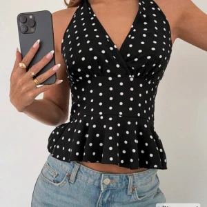 Svart prickig halterneck topp - Svart halterneck topp med vita prickar och volang nertill. Toppen har omlott-skärning framtill och knyts i nacken. Perfekt för dig som gillar retrostil och vill ha något som sticker ut.