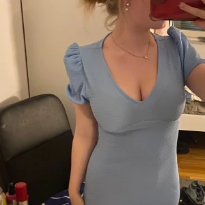 Ljusblå puffärmad klänning - Supersöt ljusblå långklänning med puffärmar och v-ringning. Klänningen är figurnära och har en kort längd, perfekt för dig som gillar en feminin och trendig stil. Materialet är lätt strukturerat och ger en snygg passform.