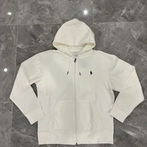 Vit hoodie från Polo Ralph Lauren - Vit hoodie från Polo Ralph Lauren med klassisk svart broderad logga på bröstet. Tröjan har huva med snörning, dragkedja framtill och två fickor. Tillverkad i mjuk bomull, perfekt för chill dagar. Snygg och enkel design som funkar till allt.