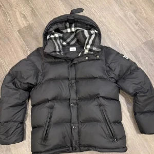 Svart pufferjacka från Burberry - Svart pufferjacka från Burberry med stor patch på ärmen och klassiskt rutigt foder i luvan. Jackan har dragkedja och knappar framtill, två fickor med dragkedja och en rejäl huva. Perfekt för kalla dagar med sin tjocka och fluffiga design.