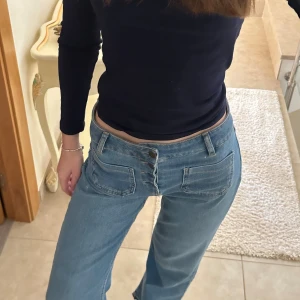 Lågmidjade blå jeans med knappar - Snygga blå jeans med låg midja och raka ben. Framtill finns stora fickor och en cool knappgylf med fyra synliga knappar. Jeansen har en avslappnad passform och är tillverkade i klassiskt denimtyg.