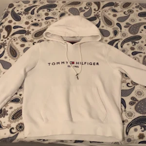 Vit hoodie från Tommy Hilfiger - Vit hoodie från Tommy Hilfiger med klassisk logga och text på bröstet. Tröjan har huva med snörning och en stor magficka. Perfekt för dig som gillar stilren och enkel design med en touch av streetwear.