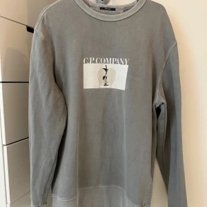 Grå sweatshirt från C.P. Company - Snygg grå sweatshirt från C.P. Company med tryck av äpple och logga både fram och bak. Tröjan har rund hals och långa ärmar, perfekt för dig som gillar stilrena och coola plagg. Materialet är mjukt och skönt, passar till jeans eller cargo. Inga defekter.