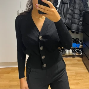 Svart kofta med stora knappar - Snygg svart kofta med v-ringning och stora, silvriga knappar framtill. Modellen är långärmad och har en normal passform som sitter snyggt i midjan. Perfekt att styla med dina favoritjeans eller kostymbyxor för en clean look.