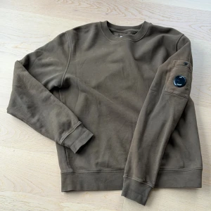 Olivgrön sweatshirt från C.P. Company - Säljer en brun sweatshirt från C.P. Company med deras ikoniska linsdetalj på ärmen. Tröjan har rund hals, ribbade muddar och en ficka med knapp på vänster ärm. Något solblekt på ställen, syns bara i vissa ljus. Lite flammig. 