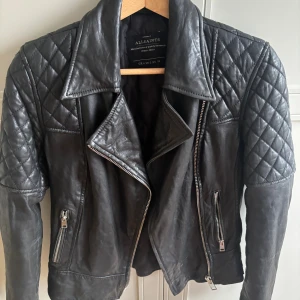 Svart skinnjacka AllSaints quiltad - Snygg svart skinnjacka från AllSaints med quiltade axlar och ärmar, coola dragkedjor och spännen i midjan. Jackan har klassisk bikerstil med bred krage och detaljer i silver. Perfekt för dig som vill ha en edgy look. Knappt använt köpt i London.