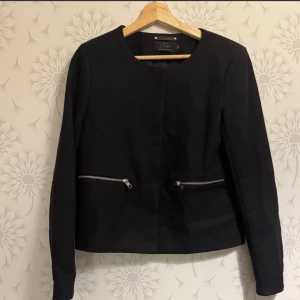 Svart kavaj från ONLY med dragkedjor - Stilren svart kavaj från ONLY i storlek 38. Jackan har rund halsringning, långa ärmar och två snygga dragkedjor framtill. Materialet är strukturerat och ger en modern känsla. Perfekt för dig som gillar en clean look med edge.