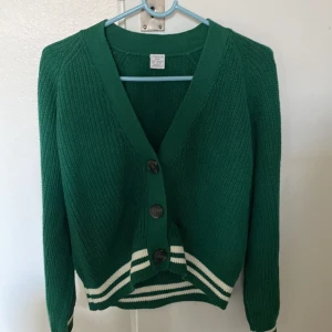 Grön kofta från Lindex med knappar - Grön ribbstickad kofta från Lindex med v-ringning och tre stora knappar framtill. Nederkanten och ärmsluten har vita ränder som ger en  elegant vibe. Koftan är långärmad och har en mjuk, stickad känsla som passar perfekt till en vardag. 