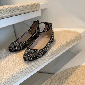 Svarta ballerinaskor med nitar från Zara - Svarta ballerinaskor från Zara med rund tå, dekorerade med silvriga nitar och små glittrande stenar över hela skon. Skorna har en låg klack och smal ankelrem med spänne. 