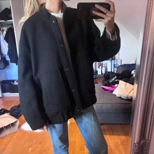 Svart oversized bomberjacka i ull - Säljer en svart bomberjacka med oversized passform med en lite högre krage. Jackan har stora fickor framtill och knäppning med tryckknappar. Materialet är tjock ull.