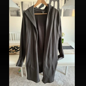 Mörkgrå cardigan jacka med huva H&M - Snygg mörkgrå cardigan jacka från H&M Divided med huva och långa ärmar. Jackan är tillverkad i mjuk bomull och har två fickor framtill. Perfekt för chill dagar och enkel att slänga på över en t-shirt eller hoodie.