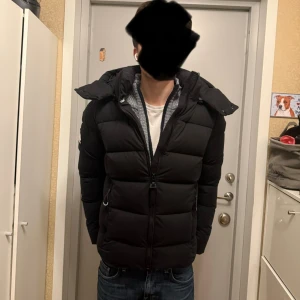 Superdry Pufferjacka - Svart pufferjacka från Superdry med huva och tydlig logga på ärmen. Jackan har dragkedja framtill, två sidofickor med dragkedja. Perfekt för kalla dagar.