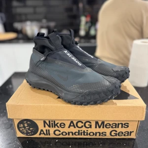 Nike ACG GORE-TEX svarta sneakers - Nike ACG sneakers med GORE-TEX, perfekta för alla väder. Svart och grå design med robust mönstrad sula för extra grepp. Snygga detaljer med ACG-logga bak och dragflik vid skaftet. Vattentätt material och sportig look, passar dig som gillar äventyr och streetstyle.