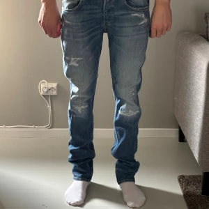Replay ljusblå slitna jeans - Säljer ett par ljusblå jeans från Replay med slitna detaljer och snygg tvätt. Klassisk femficksmodell med raka ben och normal passform. Jeansen har läderpatch med Replay-logga bak i midjan och är tillverkade i mjukt jeanstyg.