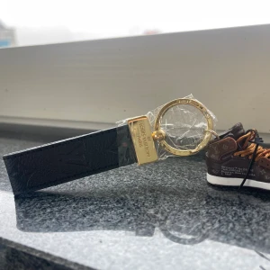  Vuitton nyckelring med sneaker - Exklusiv  Vuitton nyckelring med svart skinnrem och guldfärgad metallring. Hängande miniatyrsneaker i brunt med klassiskt LV-mönster och orange detaljer. Perfekt accessoar för dig som gillar streetstyle och lyxiga detaljer.