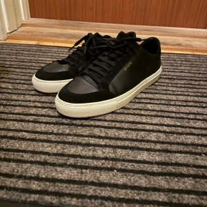 Svarta sneakers från Axel Arigato - Snygga svarta sneakers från Axel Arigato med vit platt sula och diskret guldfärgad logga på sidan. Skorna har snörning och är tillverkade i skinn och mocka för en stilren look. Perfekta för dig som gillar minimalistisk design.