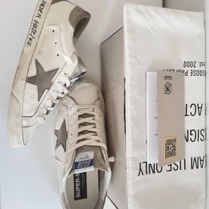 Golden Goose Superstar sneakers vit/grå - Golden Goose Superstar sneakers i vitt skinn med grå mockadetaljer och ikonisk stjärna på sidan. Klassisk låg modell med snörning, grå häl och platt gummisula. Kommer med originalkartong och dustbag. Perfekta för en cool och avslappnad stil. Obs! Pris kan diskuteras.