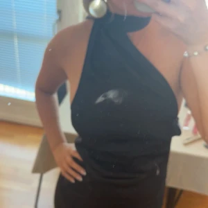 Svart långklänning - Säljer denna svarta långklänningen i satin med halterneck och oneshoulder-design. Den har dragkedja i bak och går ner till marken på mig som är 167 cm. Perfekt till bal eller finsittning!