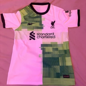 Liverpool Salah fotbollströja Nike - Liverpool FC fotbollströja med M. Salah och nummer 11 på ryggen. Tröjan är vit med gröna grafiska mönster, svarta detaljer och klubbmärke på bröstet. Tillverkad i lätt polyester, kortärmad och har sponsortryck från Standard Chartered.