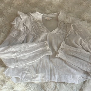 Vit blus med volanger från Gina Tricot - Vit blus från Gina Tricot med v-ringning, volanger på axlarna och spetsdetaljer. Blusen är långärmad och har lätt puffade ärmar. 