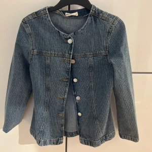 Blå jeansjacka med rund hals - Snygg blå jeansjacka från Other Stories med rund halsringning och raka långa ärmar. Jackan har framknäppning med silvriga knappar och klassiska sömmar som ger en cool vintagekänsla. Perfekt att styla med dina favoritjeans eller över en hoodie.