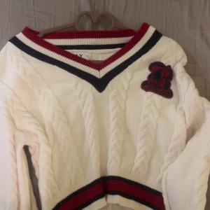 Vit kabelstickad tröja med v-ringning - Kabelstickad vit tröja med v-ringad hals och breda ribbade kanter i rött och marinblått. Broderad patch på bröstet.