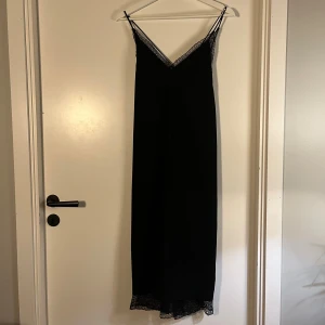 Svart långklänning med spets från H&M - Svart långklänning från H&M med tunna axelband och v-ringad hals och rygg. Klänningen har spetsdetaljer vid halsringningen, rygg och längst ner vid fållen, vilket ger en elegant touch. Materialet är mjukt och lätt, perfekt för dig som gillar stilrena och klassiska looks.