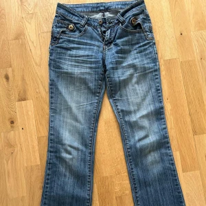 Blå jeans med broderad kant och hjärtan - Snygga blå jeans med bootcut passform och unika broderade detaljer längst ner på benen. Brodyren har hjärtan, stjärnor och ränder i rött, vitt och blått. Jeansen har coola fickor med knappar och kontrastsömmar. Perfekta för dig som vill sticka ut lite extra.