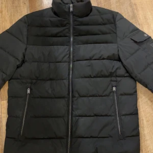 Svart pufferjacka från BOSS - Svart pufferjacka från BOSS med stilren design och raka linjer. Jackan har hög krage, två dragkedjeförsedda fickor framtill och en ärmficka med BOSS-logga. Tillverkad i syntetmaterial och perfekt för kyliga dagar. Står inte storlek men sitter som m en som bär S skulle också kunna ha den