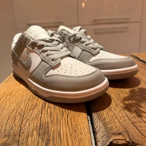 Nike Dunk Low grey fog - Snygga Nike Dunk Low sneakers i en clean färgkombination av ljusgrått och vitt. Skorna har klassisk siluett med rund tå, snörning och platt sula. Ovandelen är i skinn och syntet, med ikoniska swoosh-loggan på sidorna och broderad Nike-text på hälen.