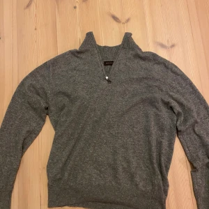 Grå kashmir half zip från Oscar Jacobson - Snygg grå kashmir tröja från Oscar Jacobson med half zip och hög krage. Tröjan har långärmade ribbade muddar och är tillverkad i mjukt material som passar perfekt till jeans. Enkel och stilren design som funkar året runt.