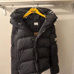 Burberry dunjacka  - Exklusiv Burberry London England dunjacka i mörkgrå färg, i mycket fint skick. Klassisk design med rutigt luvfoder och logotyp på ärmen. Ärmarna kan tas av så jackan blir till en stilren väst och perfekt året runt. Varm, lätt och elegant