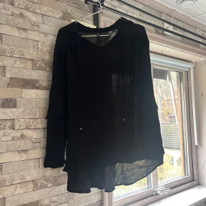 Svart transparent blus  - Svart transparent blus i storlek XL. Blusen har långa ärmar, v-ringning och tunna knytband vid halsen. Den är lätt och luftig med en något längre bakdel, perfekt för en cool och avslappnad look.