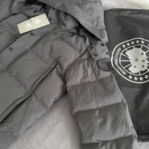 Canada Goose jacka  - Snygg grå dunjacka från Canada Goose med svart logotyp på ärmen och avtagbar huva. Jackan har raka linjer, stora fickor med dragkedja och knappar framtill. Perfekt för kalla dagar och har en klassisk, stilren look.