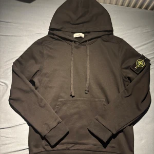 Svart hoodie från Stone Island - Svart hoodie från Stone Island med klassisk logotyp-patch på vänster ärm. Tröjan har huva med snörning, magficka och ribbade muddar. Materialet är mjuk bomull och passformen är normal. Perfekt för dig som gillar streetwear och vill ha en clean look.