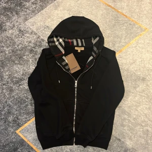 Svart Burberry hoodie med dragkedja - Svart hoodie från Burberry med klassiskt rutigt foder i huvan och snygga silverdetaljer på dragkedjan och snörningen. Tröjan har broderad Burberry-logga på bröstet och är tillverkad i mjuk bomull. Perfekt för dig som gillar stilrena och exklusiva plagg.
