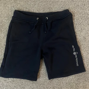 Svarta shorts från Sail Racing - Svarta shorts från Sail Racing med snörning i midjan och tryckt logga på ena benet. Mjukt material, två fickor fram och en bakficka. Perfekta för sommaren och chill dagar.