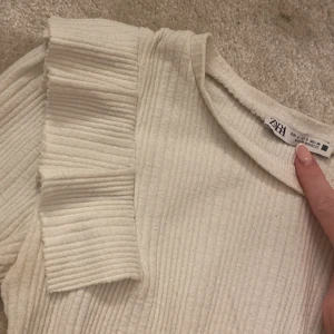 Vit ribbad topp med volang från Zara - Säljer en vit ribbad topp från Zara i storlek S. Toppen har korta ärmar och en snygg volangdetalj på axeln som ger extra edge. Materialet är mjukt och stretchigt, perfekt för dig som gillar stilrena och coola plagg. Passformen är normal och toppen har rund hals.