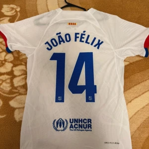 Vit FC Barcelona João Félix tröja Nike - Säljer en vit FC Barcelona fotbollströja med João Félix och nummer 14 på ryggen. Tröjan har blå detaljer, röda och blå muddar på ärmarna, klubbmärke och Nike-logga framtill samt Spotify och UNHCR-tryck. Tillverkad i lätt funktionsmaterial.